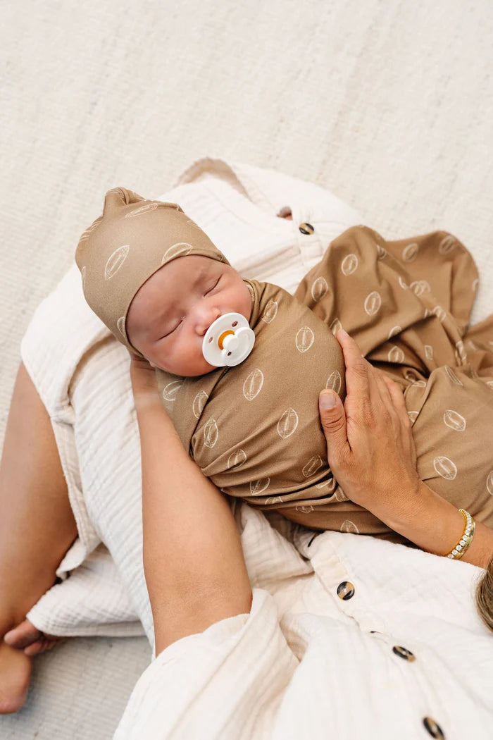 A sleeping newborn baby swaddled in a brown swaddle blanket with a matching hat, resting on a mother's lap. The swaddle and hat feature a light beige football pattern.