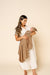 A woman in a white button-front dress holds a baby wrapped in a taupe patterned blanket against a solid beige studio background.