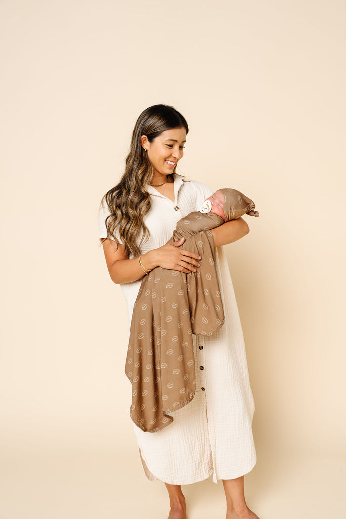 A woman in a white button-front dress holds a baby wrapped in a taupe patterned blanket against a solid beige studio background.