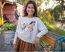 Young girl wearing a sweater with a horse design, standing outdoors.