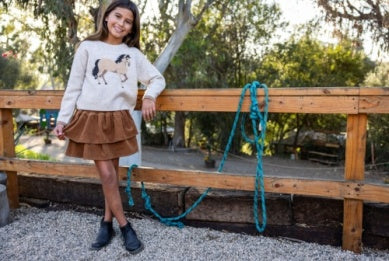 Girl wearing a sweater with a horse design, standing on a wooden platform with a rope.