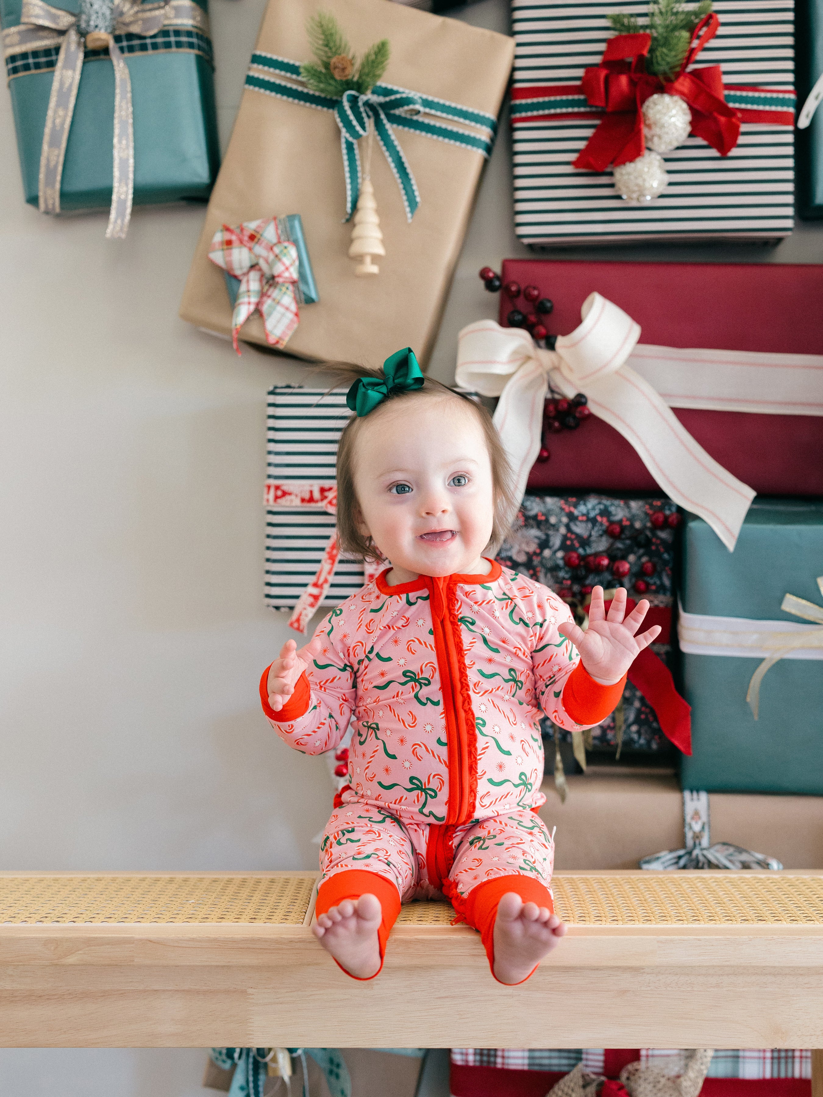 A baby wearing a pink Christmas-themed footed sleeper sits on a wooden bench in front of wrapped holiday gifts. The sleeper has a red-orange collar, cuffs, and zipper, with a pink background patterned with green pine trees and red ornaments.