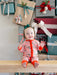 A baby wearing a pink Christmas-themed footed sleeper sits on a wooden bench in front of wrapped holiday gifts. The sleeper has a red-orange collar, cuffs, and zipper, with a pink background patterned with green pine trees and red ornaments.