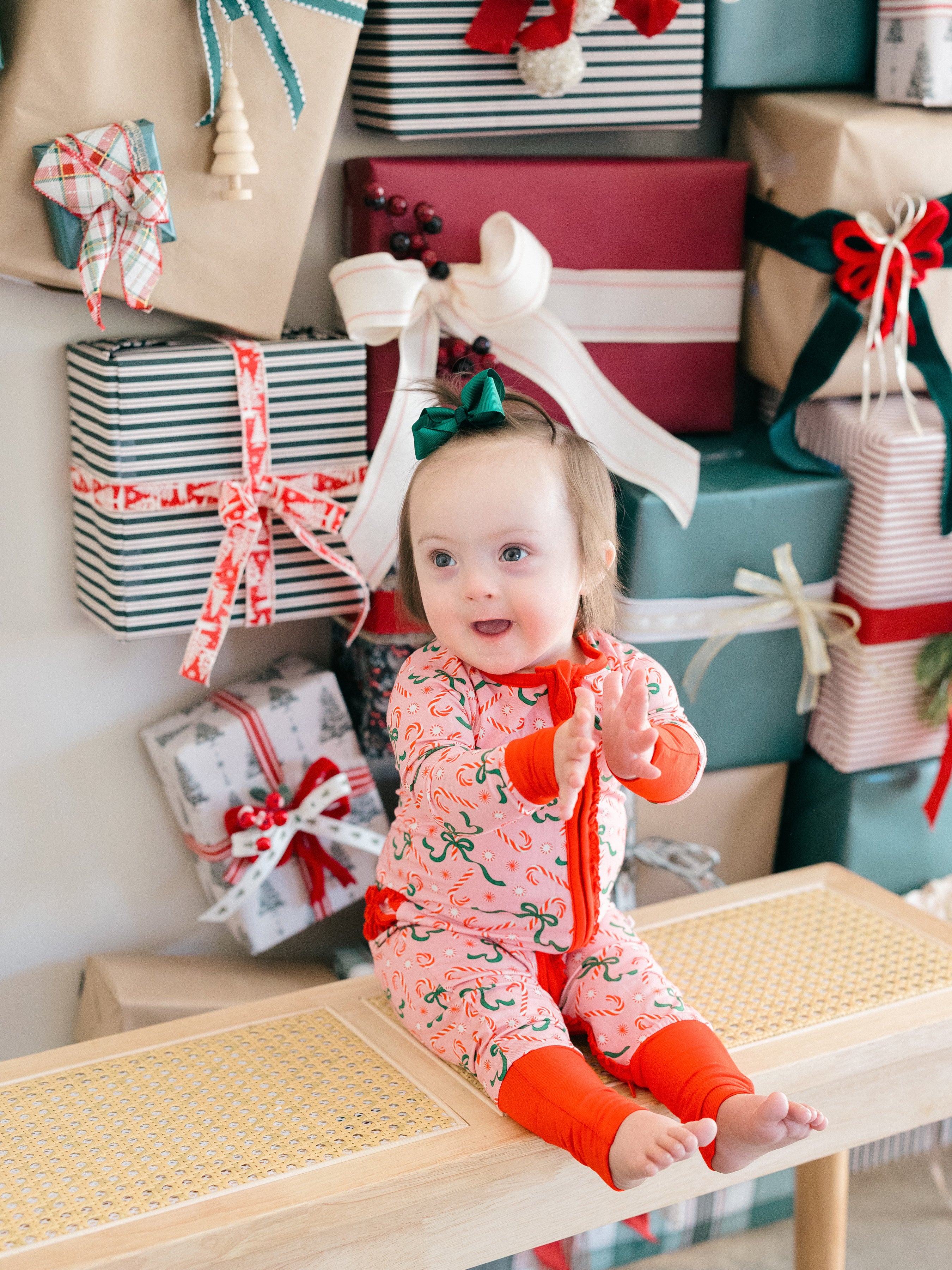 A baby wearing a festive Christmas-themed onesie with red leg warmers sits on a wooden bench surrounded by wrapped holiday presents.