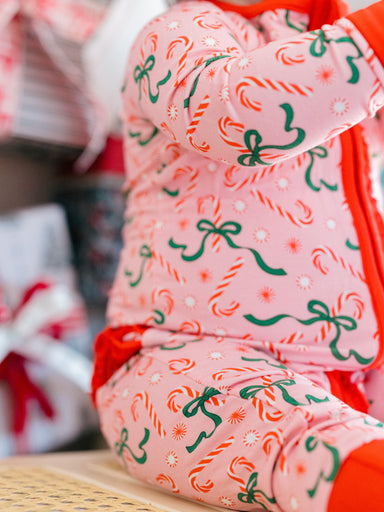 A close-up of a baby wearing a pink one-piece pajama with a festive holiday print of candy canes, green bows, and white snowflakes, paired with a bright red blanket.
