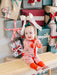 A baby wearing a festive Christmas-themed onesie with red leg warmers sits on a wooden bench surrounded by wrapped holiday presents.