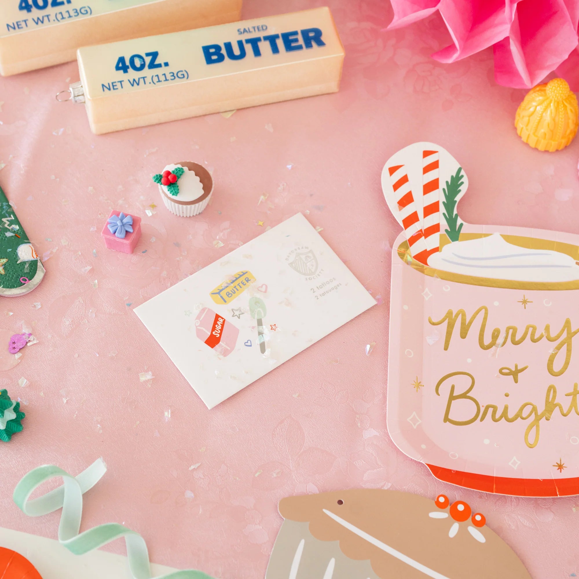 Decorative items including a block of butter, a mug with 'Merry & Bright' text, and small festive elements on a pink background.