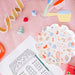 A styled flat-lay of a white scalloped paper plate printed with colorful baking and kitchen-themed illustrations, placed on a pink surface alongside party props, a coloring sheet, and holiday decorations.