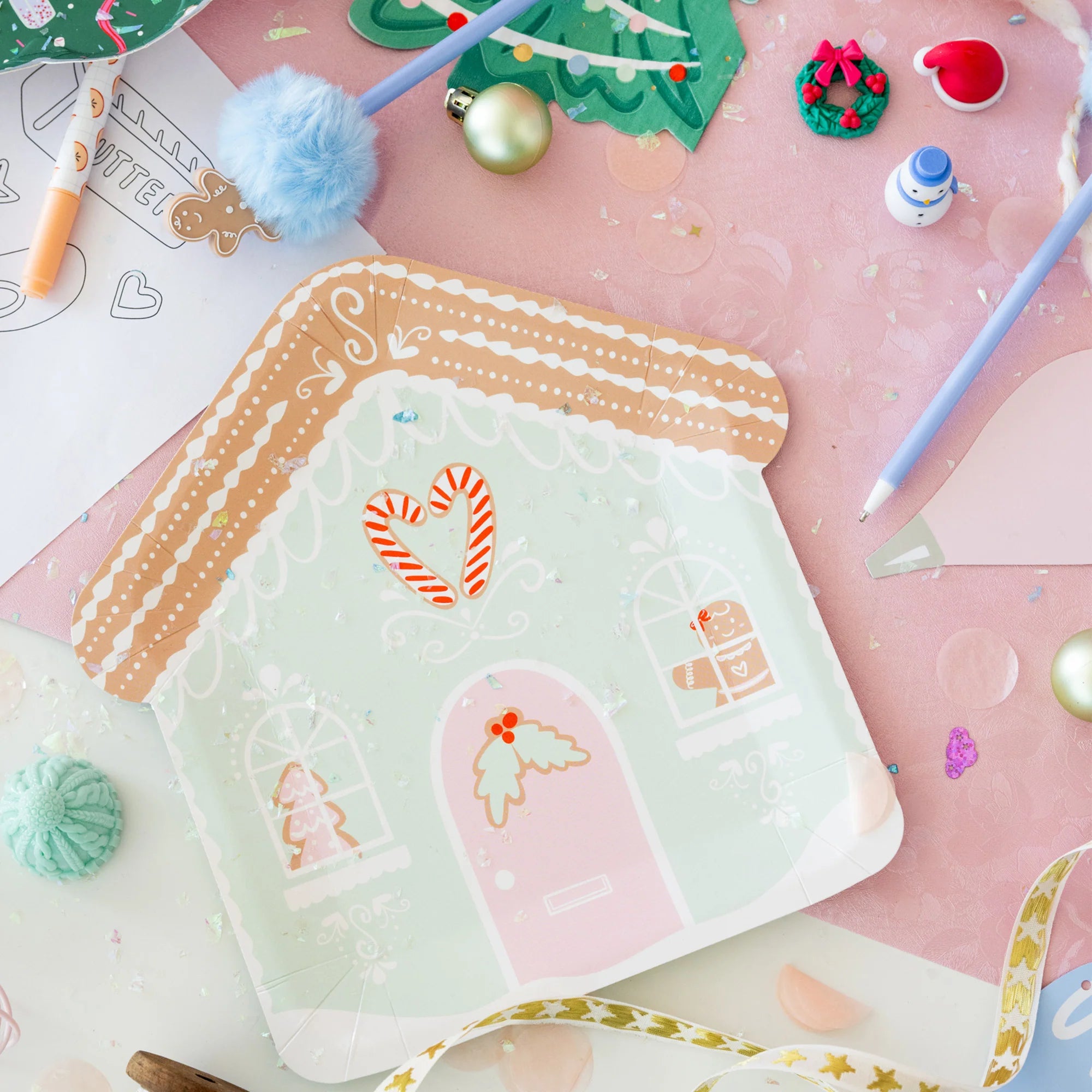 A house-shaped paper plate with a gingerbread house illustration, photographed in a styled flat-lay with holiday decorations, pens, and craft supplies on a pink surface.