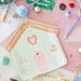 A house-shaped paper plate with a gingerbread house illustration, photographed in a styled flat-lay with holiday decorations, pens, and craft supplies on a pink surface.