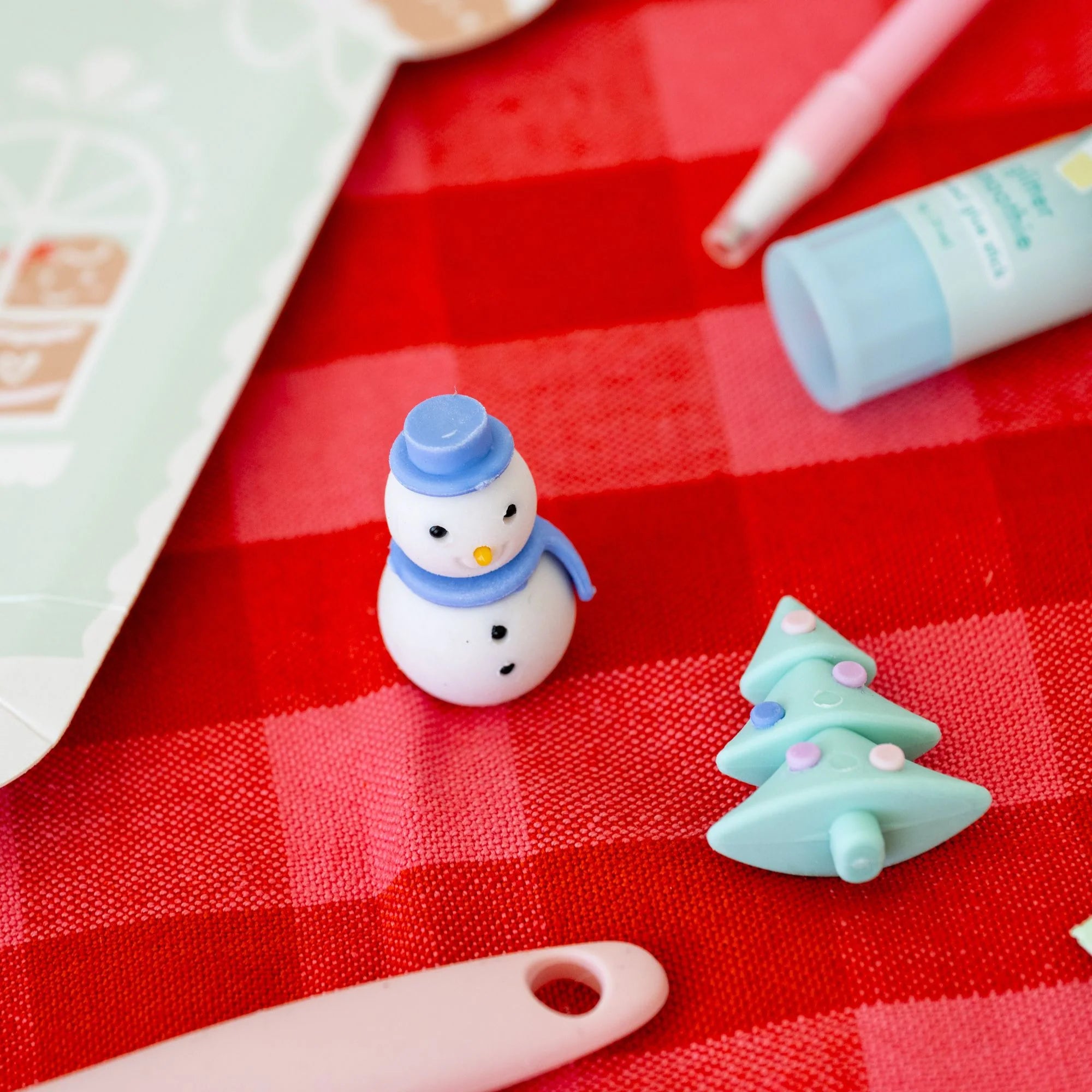 A small snowman charm and a mint green Christmas tree charm resting on a red and white checkered fabric surface, surrounded by pastel-colored stationery items.