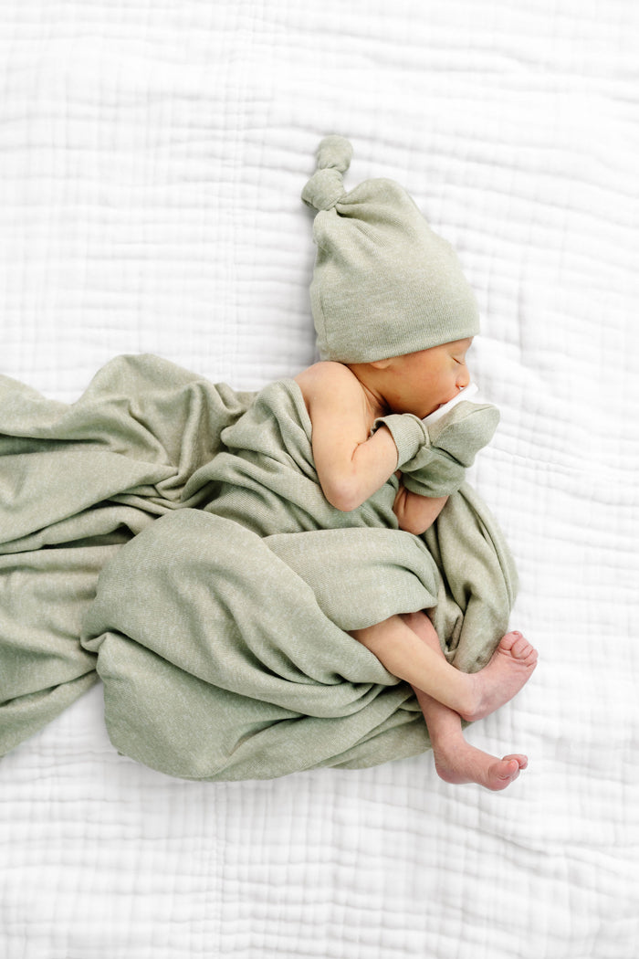 A baby laying with  a light sage green heathered fabric, including a square blanket, a knotted beanie hat, and a pair of mittens, arranged on a plain white background.