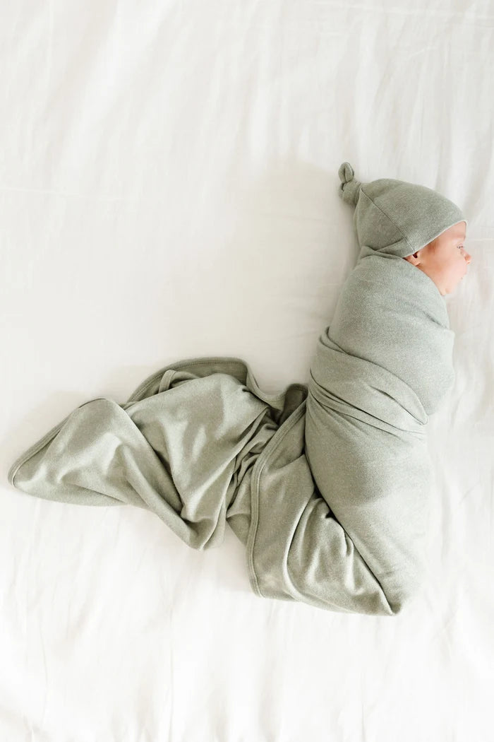 A babyswaddled in a light sage green heathered fabric, including a square blanket and a knotted beanie hat, arranged on a plain white background.