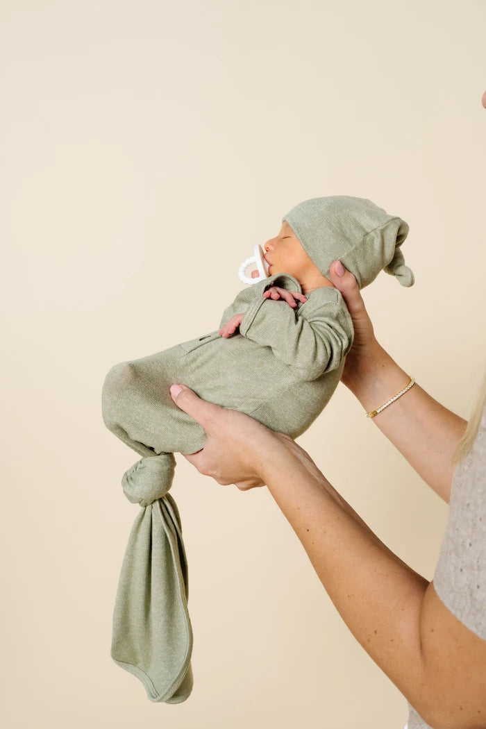 A newborn baby wearing an olive green swaddle blanket and matching beanie hat, held by an adult's hands against a solid beige background.