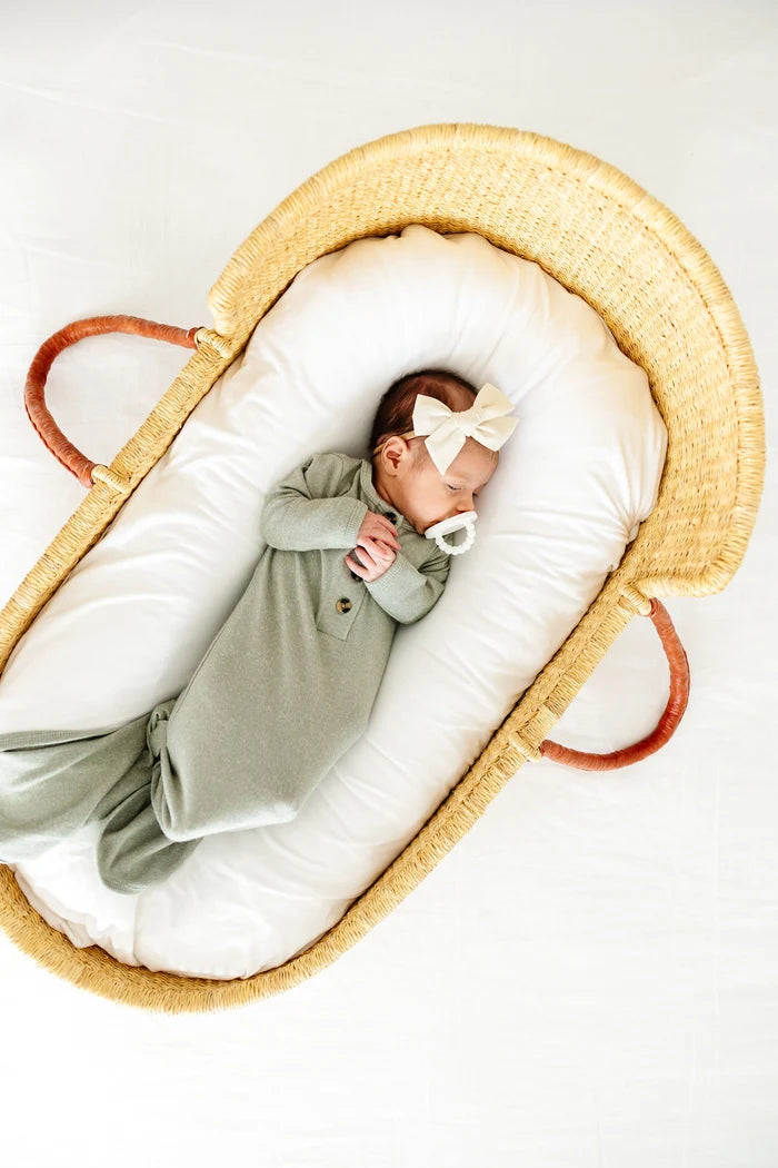 A newborn baby wearing a sage green footed sleeper and a white bow headband, sleeping inside a woven natural fiber Moses basket with white cushions.
