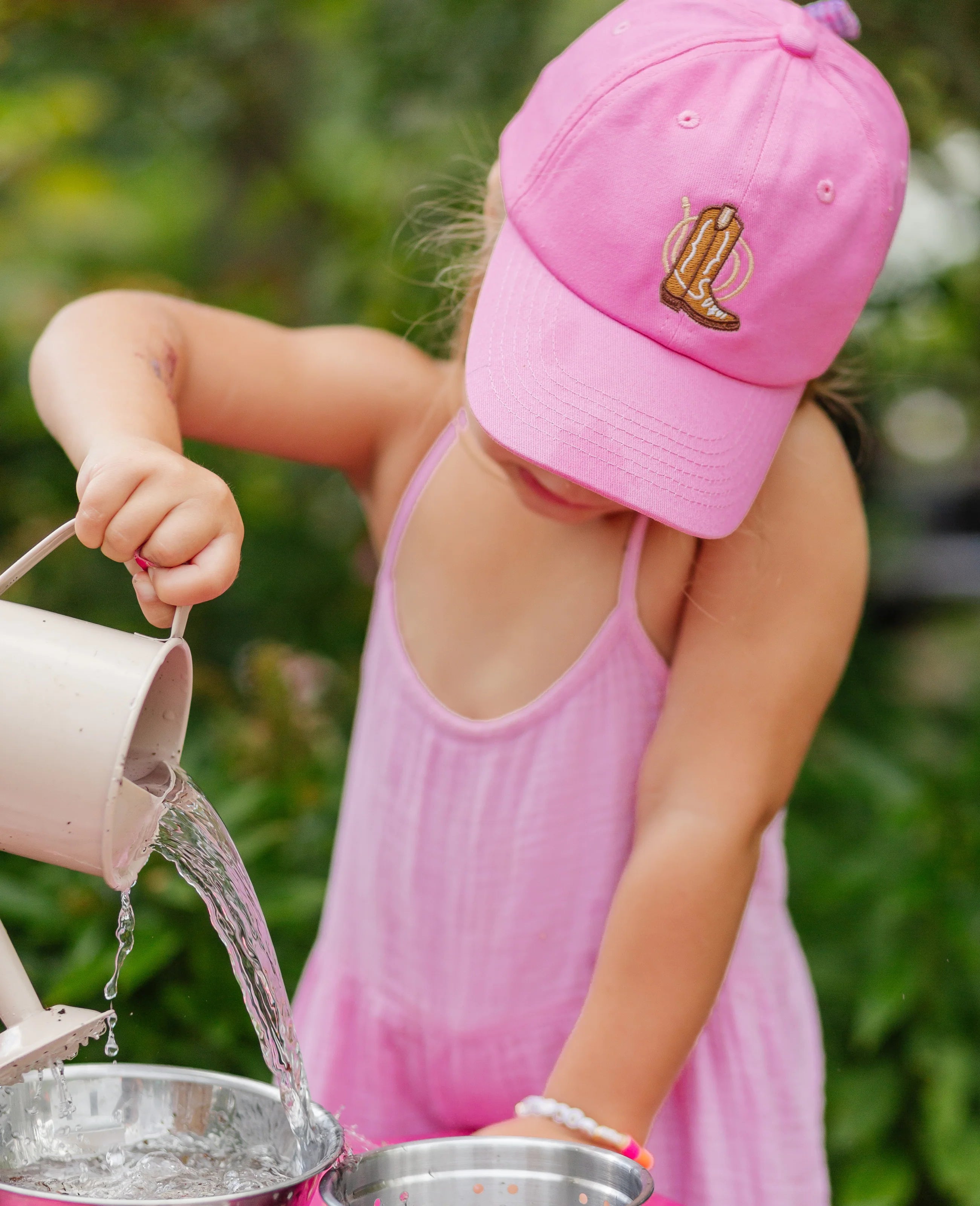 Child in pink outfit and cap pouring water from a can into another container outdoors.