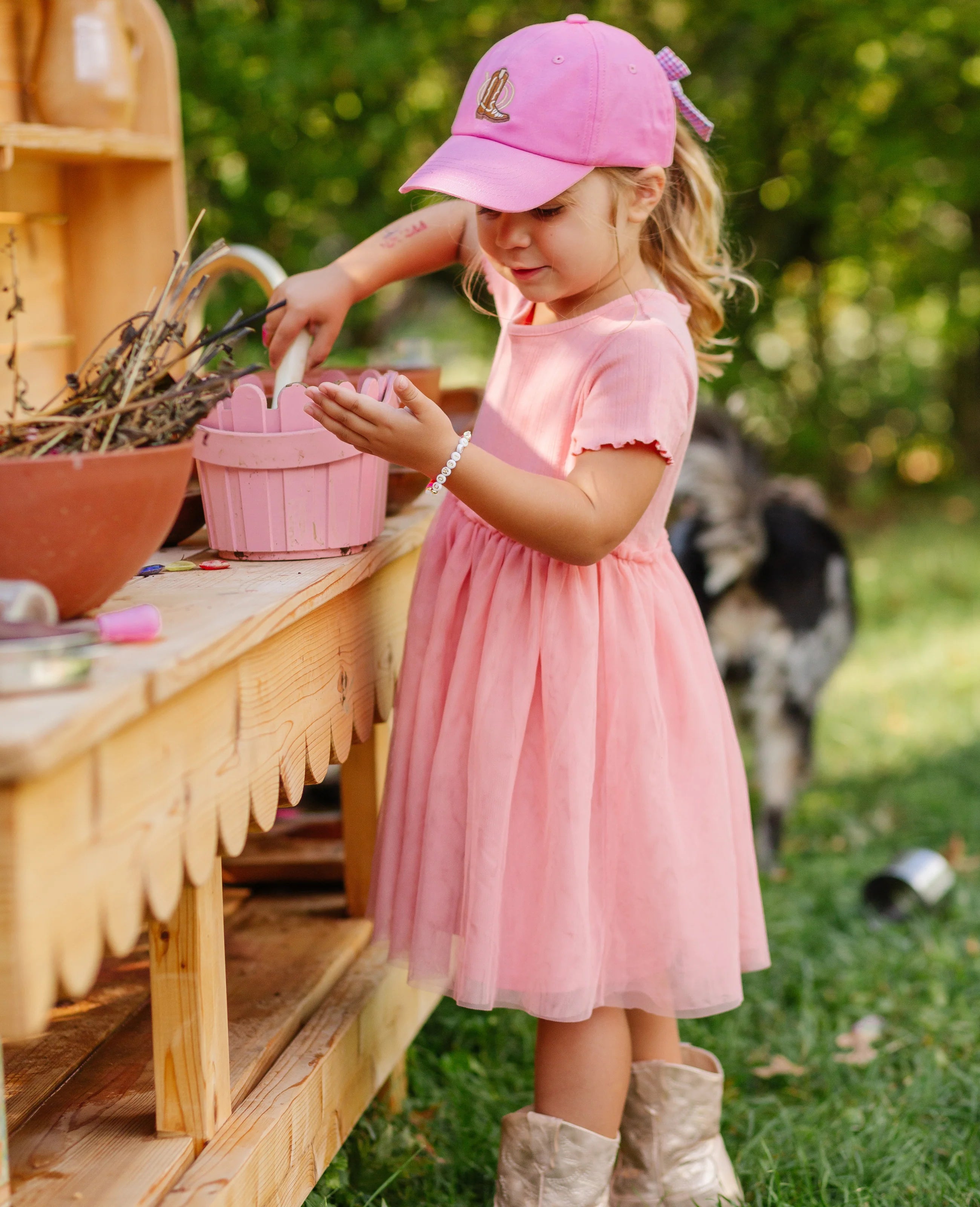 Young girl in a pink dress and cap standing outdoors with a pink basket.