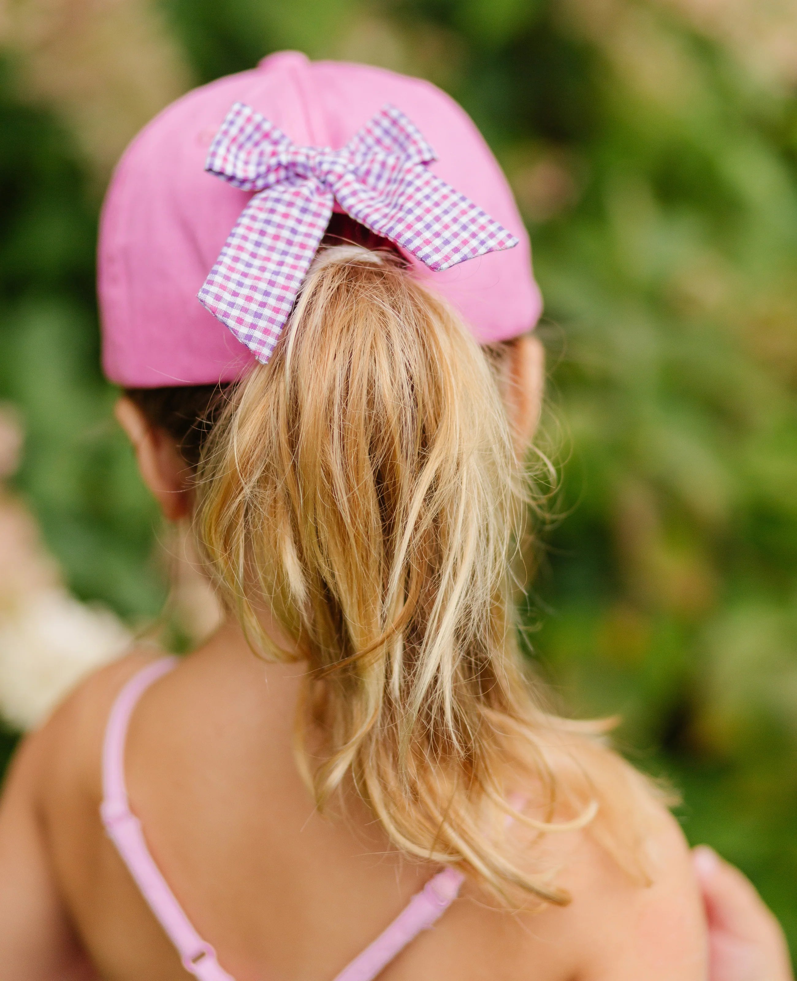Person wearing a pink cap with a large bow, standing outdoors with greenery in the background.
