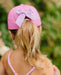 Person wearing a pink cap with a large bow, standing outdoors with greenery in the background.