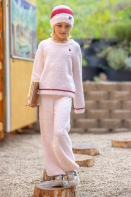 A young girl wearing a fuzzy light pink two-piece lounge set with a matching Santa hat, standing on a wooden log in an outdoor play area.
