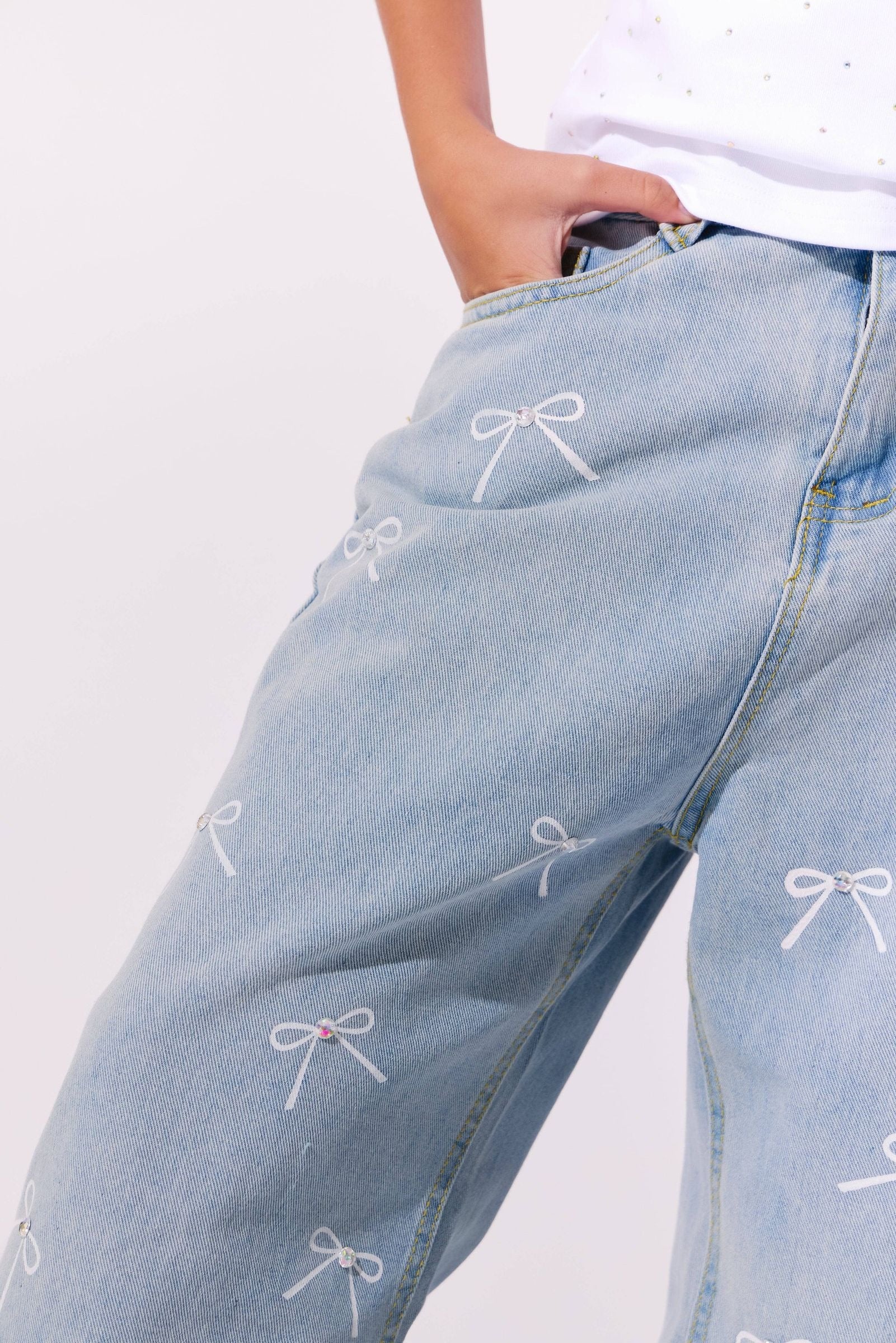 Close-up of a person wearing light blue denim jeans with white embroidered bow details, paired with a white top against a plain light background.