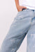 Close-up of a person wearing light blue denim jeans with white embroidered bow details, paired with a white top against a plain light background.