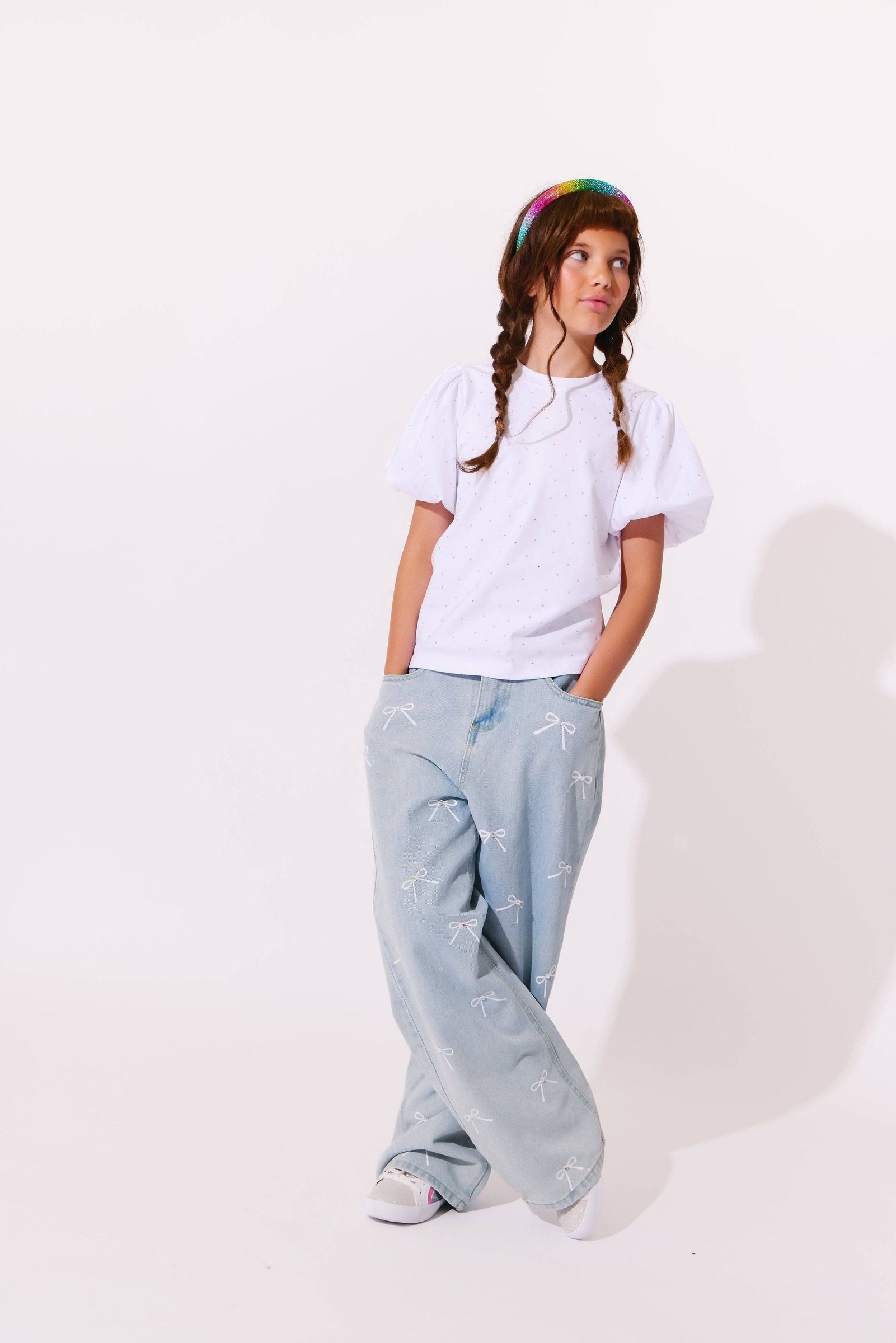 A young girl with braided hair wearing a white short-sleeve t-shirt, light blue wide-leg embroidered pants, and a rainbow-colored beanie, standing against a plain white studio background.