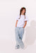 A young girl with braided hair wearing a white short-sleeve t-shirt, light blue wide-leg embroidered pants, and a rainbow-colored beanie, standing against a plain white studio background.