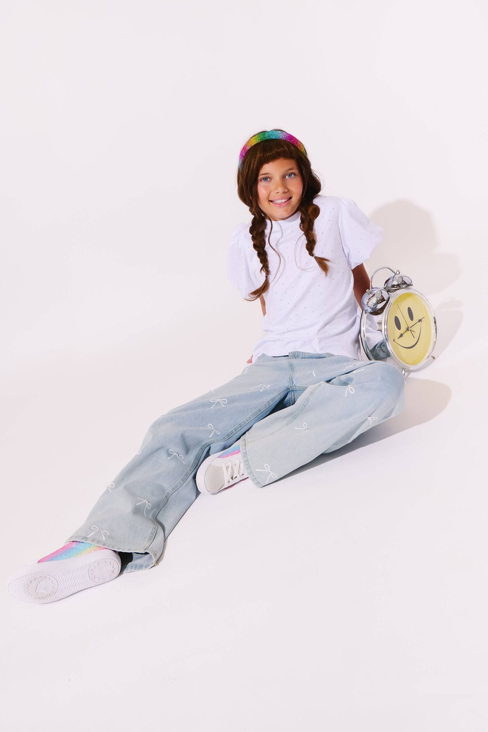 A young girl with braided hair sits on a white studio floor wearing a white t-shirt, light-wash denim jeans, white sneakers, and a rainbow beanie. She is holding a silver and yellow smiley-face backpack.