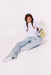 A young girl with braided hair sits on a white studio floor wearing a white t-shirt, light-wash denim jeans, white sneakers, and a rainbow beanie. She is holding a silver and yellow smiley-face backpack.
