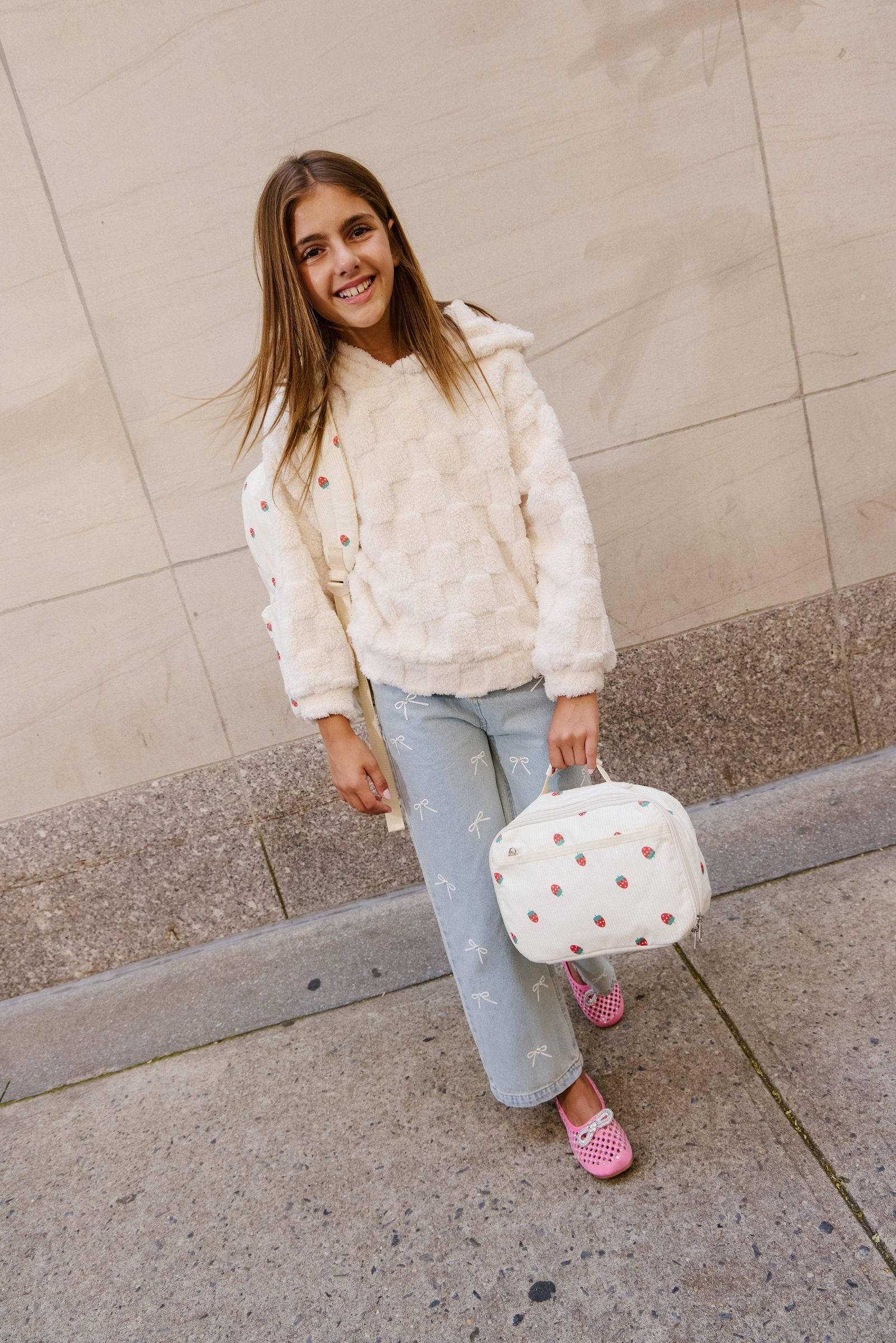 Young girl in a white fleece jacket and light blue jeans holding a polka dot bag against a beige wall.