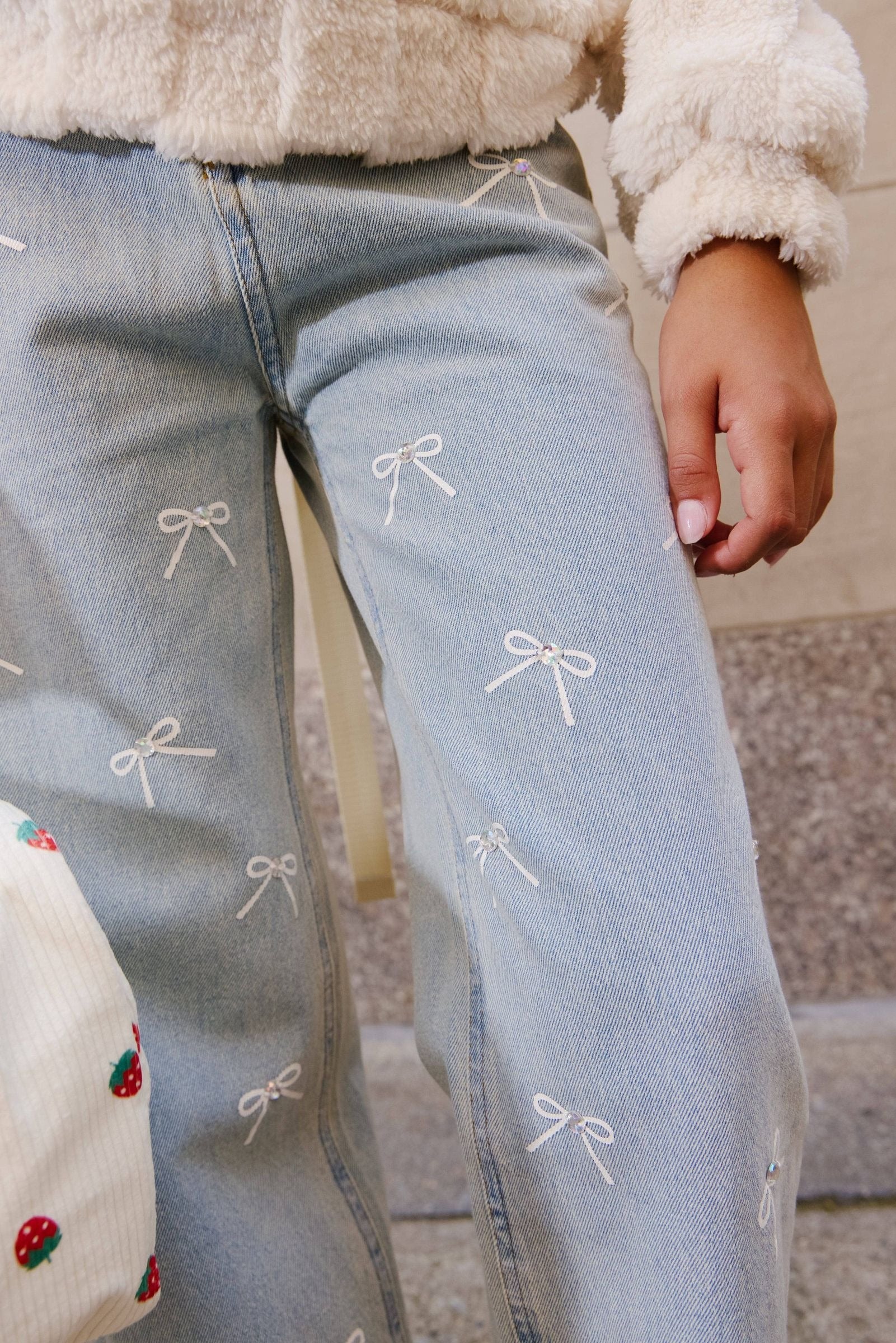 Close-up of a person wearing light blue jeans with small white embroidered bow motifs, paired with a cream fuzzy top and a strawberry-patterned bag.
