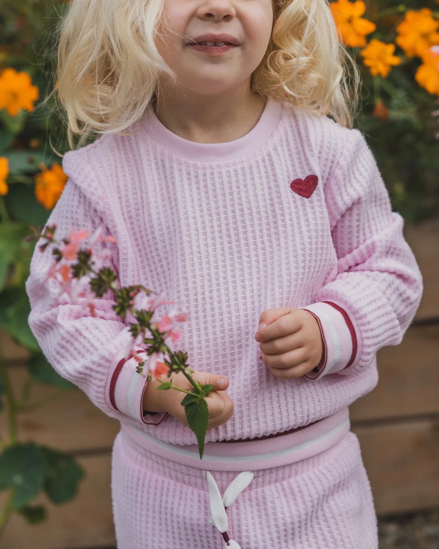 A young child wearing a light pink waffle-knit matching lounge set, featuring a sweatshirt with a small red heart on the chest and matching shorts, holding a small pink flower in a garden setting.