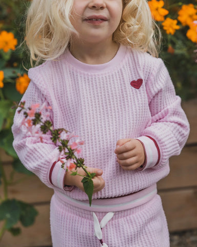A young child wearing a light pink waffle-knit matching lounge set, featuring a sweatshirt with a small red heart on the chest and matching shorts, holding a small pink flower in a garden setting.
