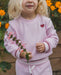 A young child wearing a light pink waffle-knit matching lounge set, featuring a sweatshirt with a small red heart on the chest and matching shorts, holding a small pink flower in a garden setting.