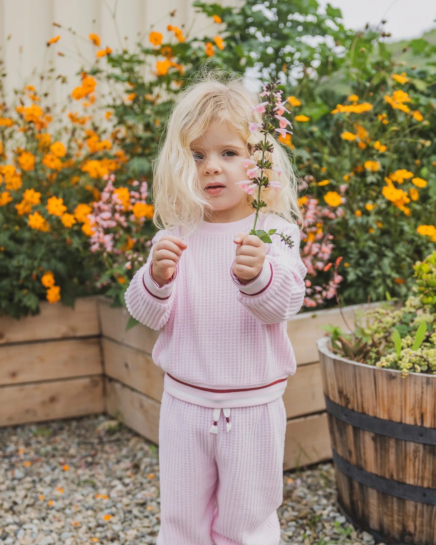 A young blonde toddler stands in a garden wearing a light pink waffle-knit matching lounge set, holding a small pink flower in front of her face. The background features wooden planter boxes filled with orange and pink flowers.