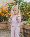 A young blonde toddler stands in a garden wearing a light pink waffle-knit matching lounge set, holding a small pink flower in front of her face. The background features wooden planter boxes filled with orange and pink flowers.