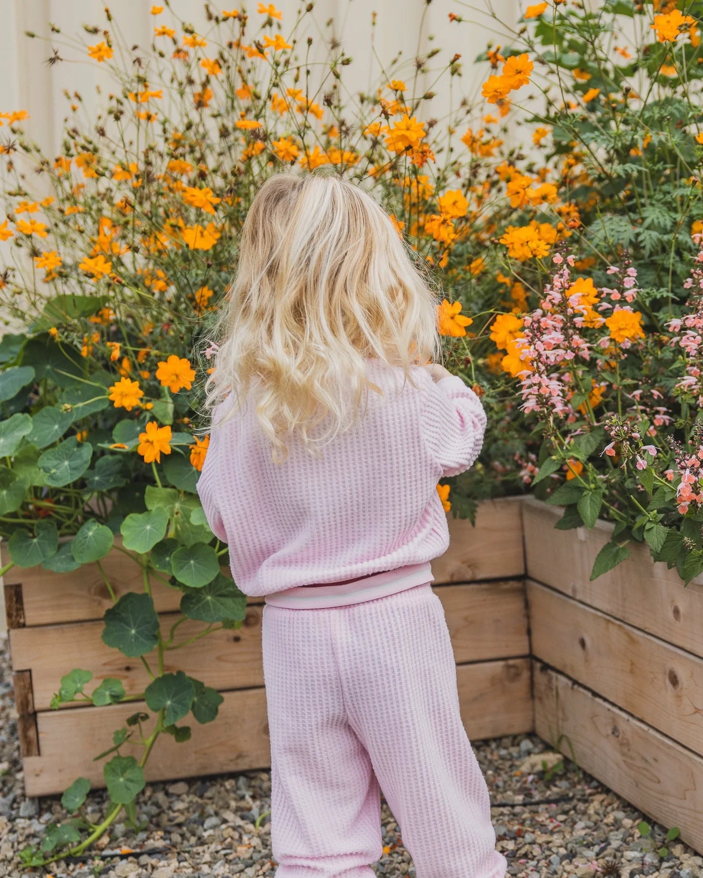 A young child with long blonde hair stands with their back to the camera, wearing a light pink waffle-knit matching lounge set in a garden setting with orange flowers and wooden planters.