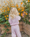 A young child with long blonde hair stands with their back to the camera, wearing a light pink waffle-knit matching lounge set in a garden setting with orange flowers and wooden planters.