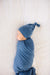 A sleeping newborn baby swaddled in a dusty blue blanket and wearing a matching blue knit hat with a small knot, lying on a white textured blanket.