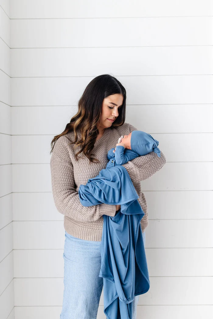 A woman in a beige knit sweater and light blue jeans holds a baby wrapped in a dusty blue blanket against a white shiplap wall.