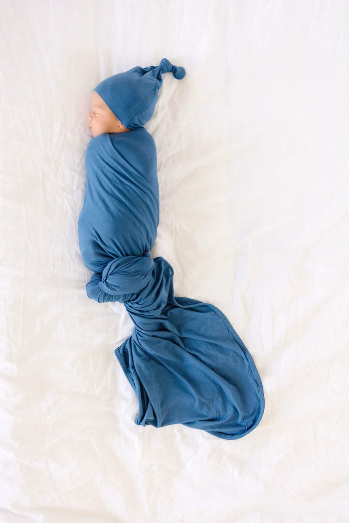A newborn baby sleeping peacefully, swaddled in a dusty blue blanket and wearing a matching blue knit hat, laid on a white textured sheet.
