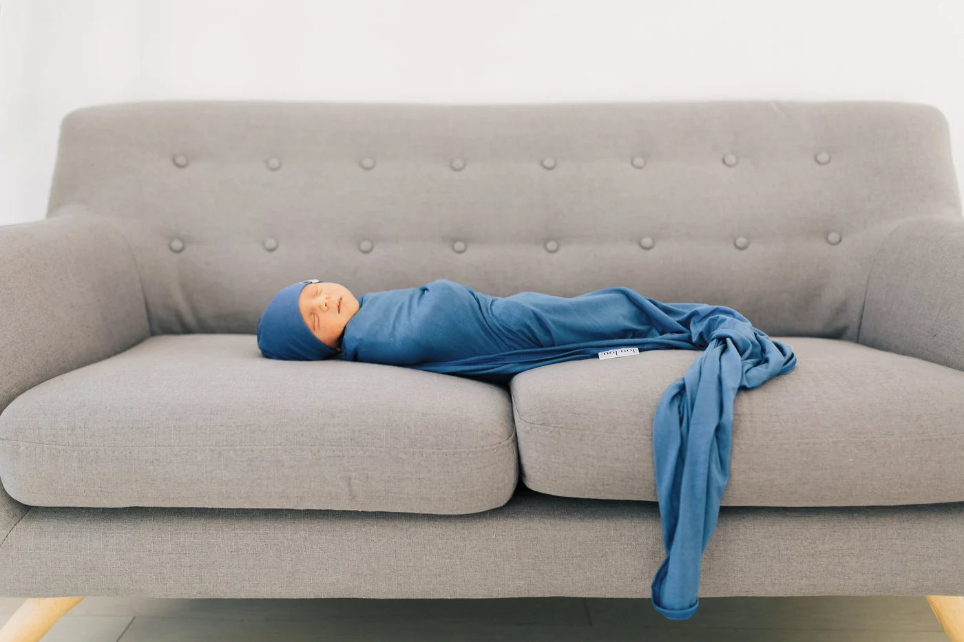 A baby swaddled in a blue blanket and wearing a matching blue hat, lying on a gray tufted sofa.