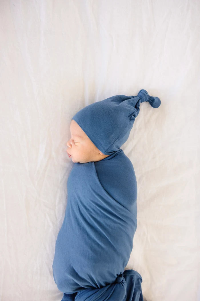 A sleeping newborn baby swaddled in a dusty blue blanket and wearing a matching blue knit hat with a small knot, lying on a white textured blanket.