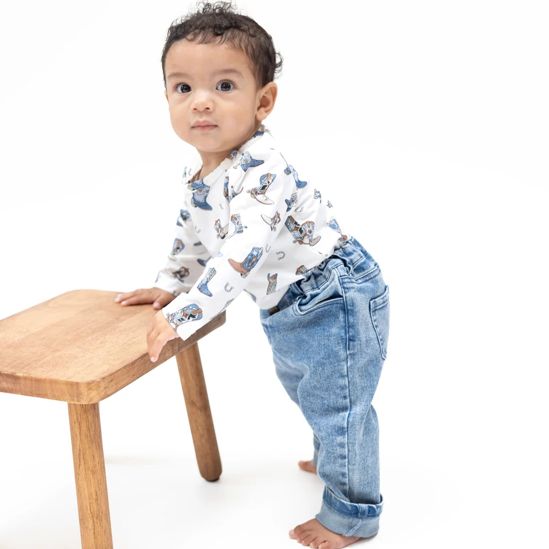 A baby toddler wearing a white long-sleeve shirt with a blue cartoon animal print and light blue jeans, standing on a white background and leaning on a small wooden stool.