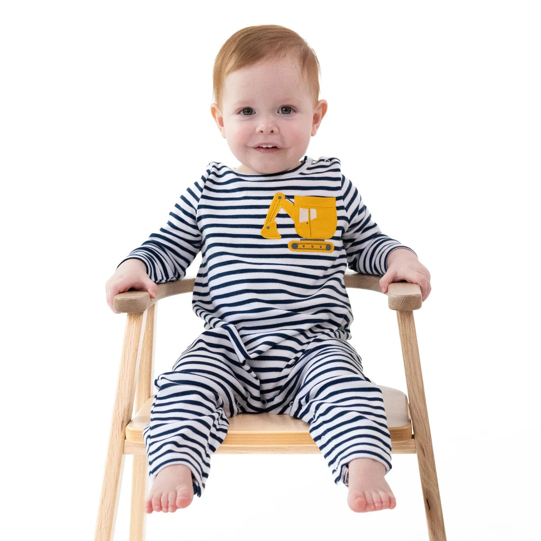 A baby sitting on a wooden chair wearing a navy and white striped long-sleeve romper with a yellow construction graphic on the chest, photographed against a solid white background.