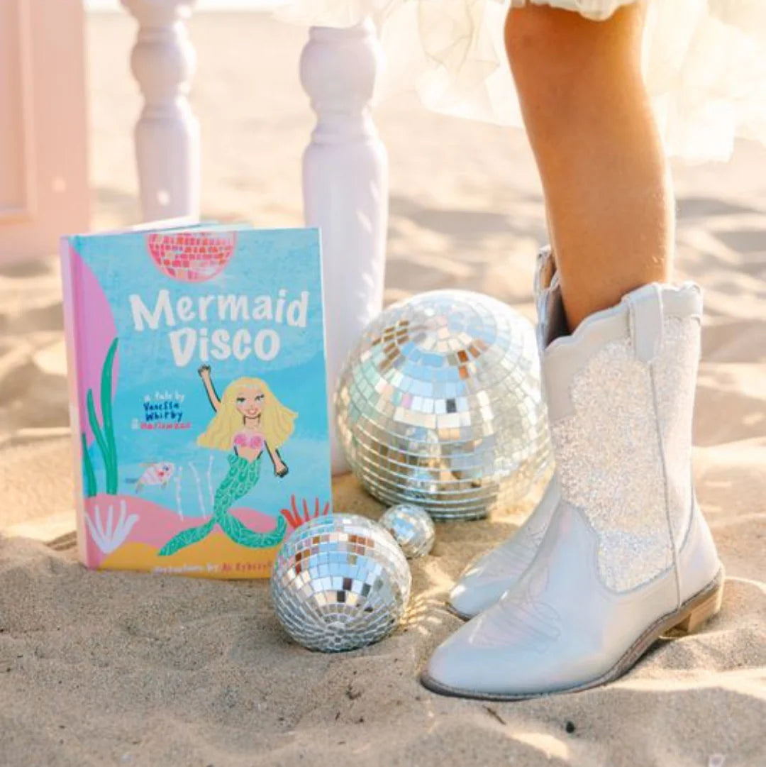Book titled 'Mermaid Disco' on sand with disco balls and white boots.