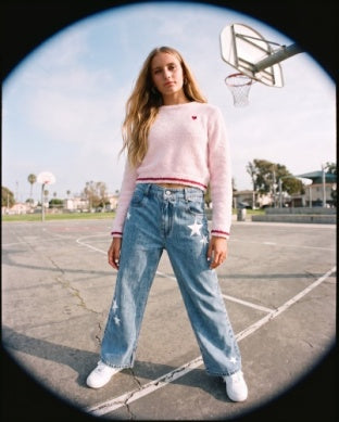 A young woman stands on an outdoor basketball court wearing a light pink long-sleeve knit sweater with a small red heart on the chest, light-wash blue jeans with white star patches, and white sneakers.