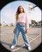 A young woman stands on an outdoor basketball court wearing a light pink long-sleeve knit sweater with a small red heart on the chest, light-wash blue jeans with white star patches, and white sneakers.
