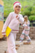 A young girl wearing a fuzzy light pink two-piece pajama set with red trim, a matching pink beanie with a red stripe, and holding a yellow notebook, standing in an outdoor playground setting.